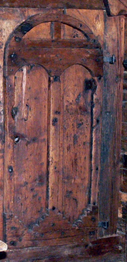 A dark, weathered plank door with an applied round-headed frame and iron studs in a log-walled Alpine room