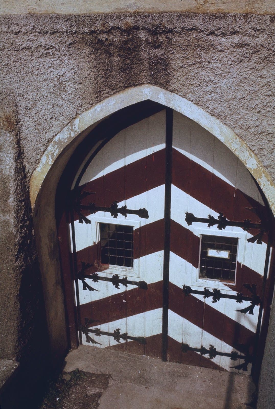 A pointed-arch wooden door painted with red and white chevrons and crossed by long black iron strap hinges