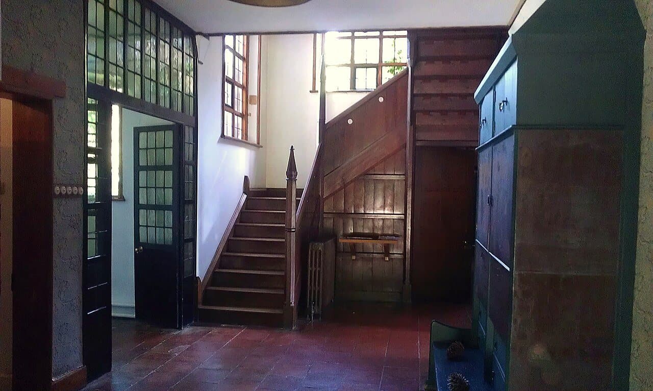 A hall with a dark oak staircase with tall pointed newel posts, leaded windows above and a large painted cupboard at the right