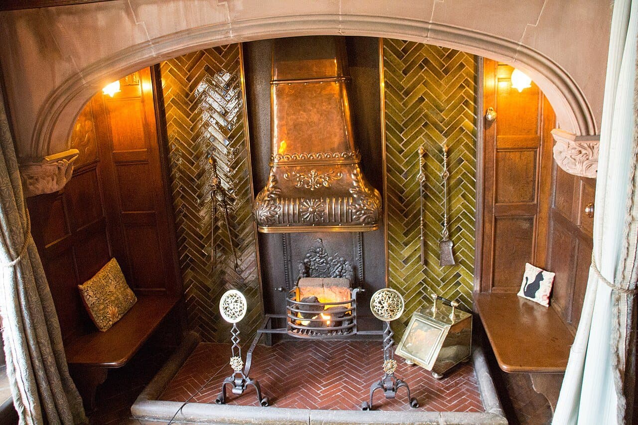 An arched inglenook with oak-paneled built-in benches on either side, green herringbone tiles and a copper hood above a fire grate