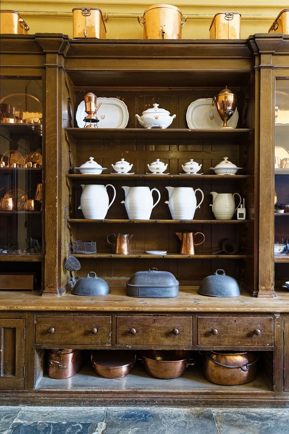 Tall wooden kitchen cupboard with shelves of white jugs, drawers and copper pans below