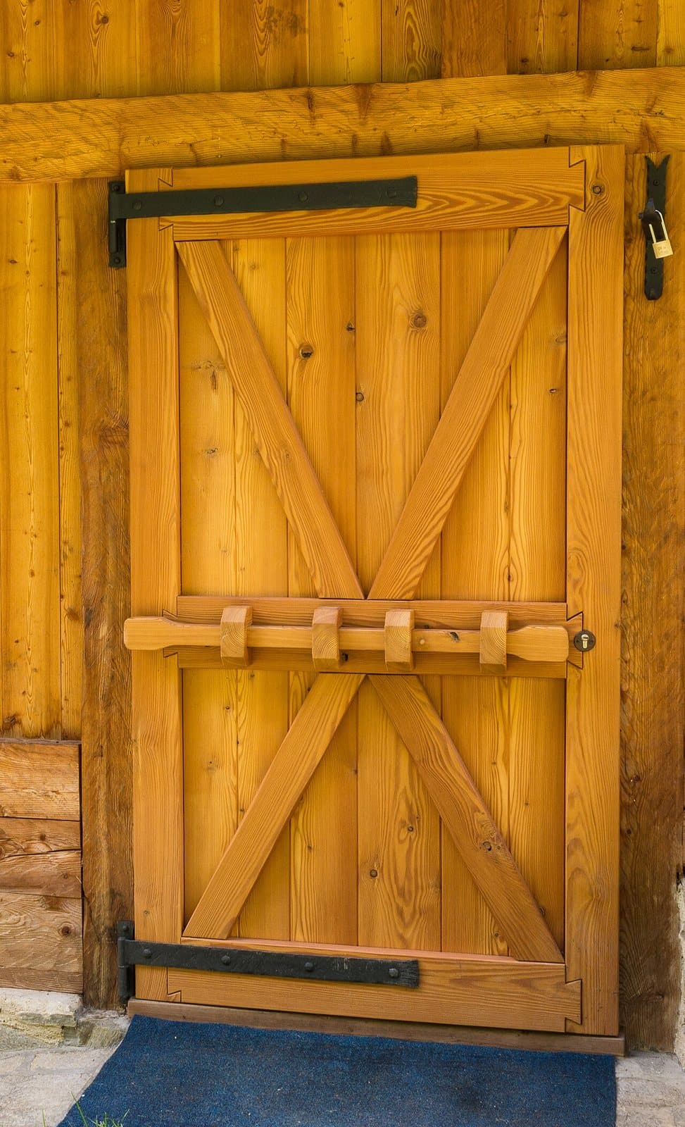 A golden wooden plank door with horizontal ledges, diagonal braces, a wooden sliding bolt and black iron strap hinges