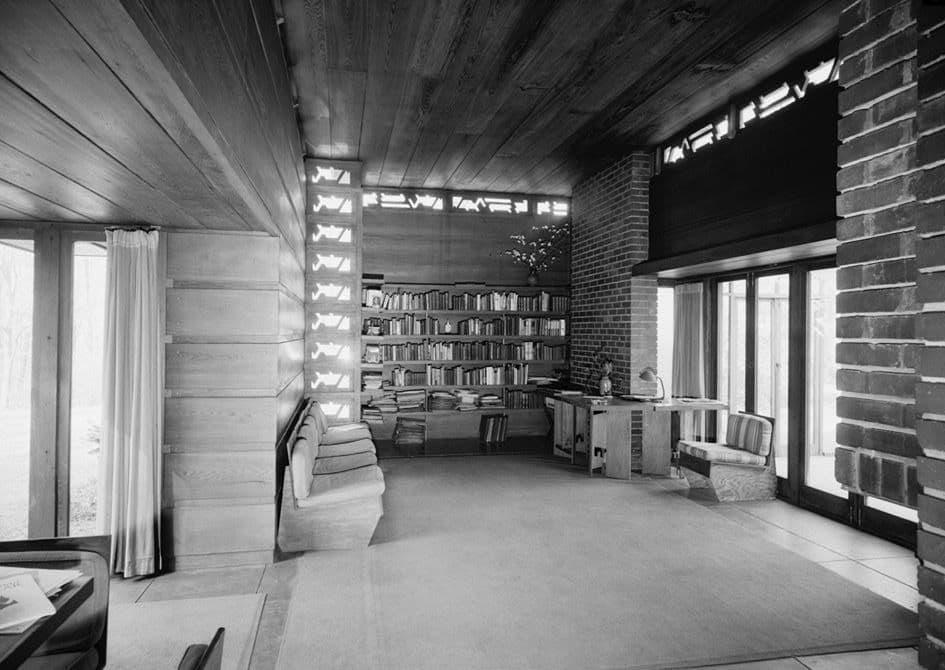Black-and-white photograph of a Usonian living room with board walls and built-in bookshelves