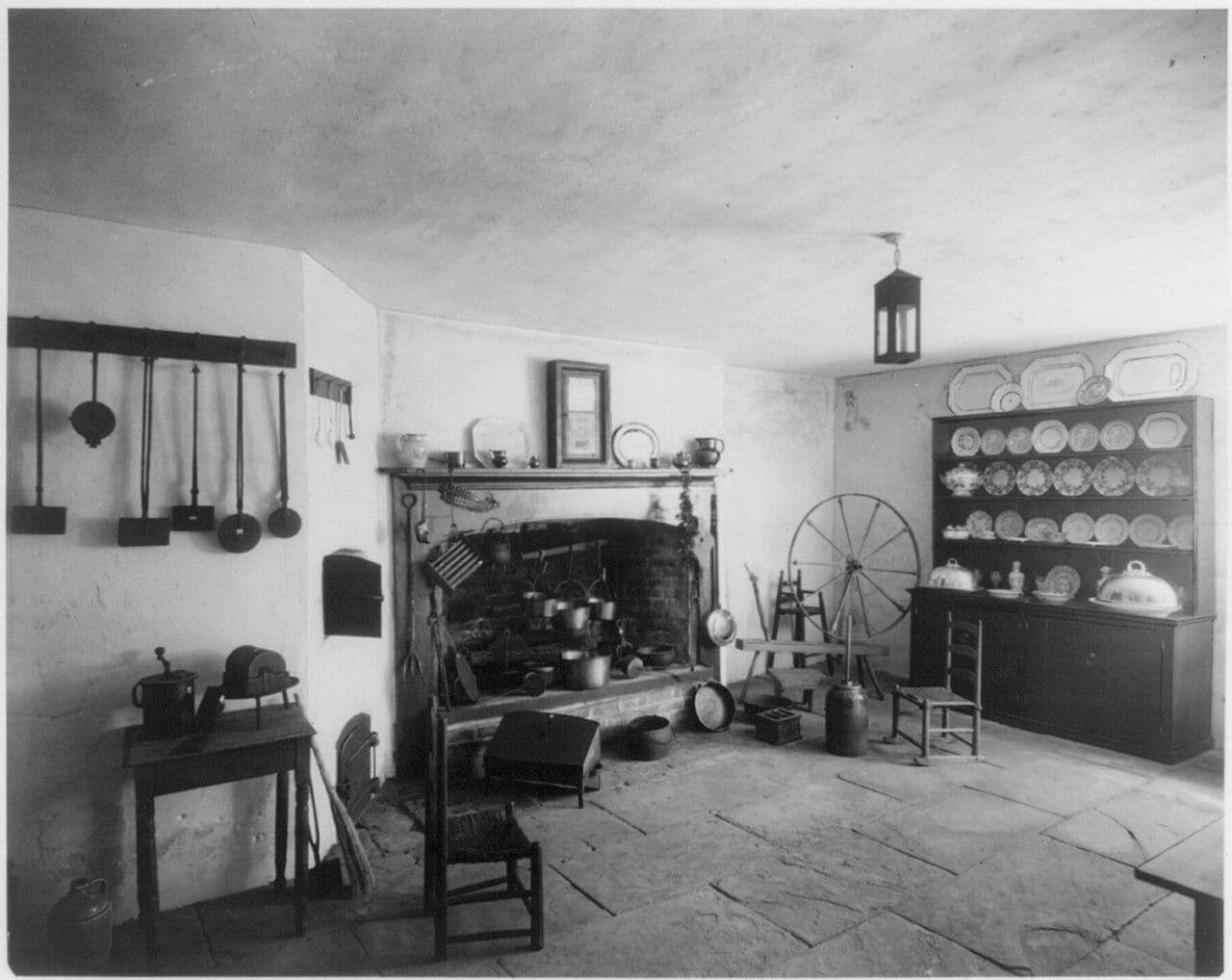 Historic photograph of an old kitchen with a large fireplace, a china hutch full of plates and a spinning wheel