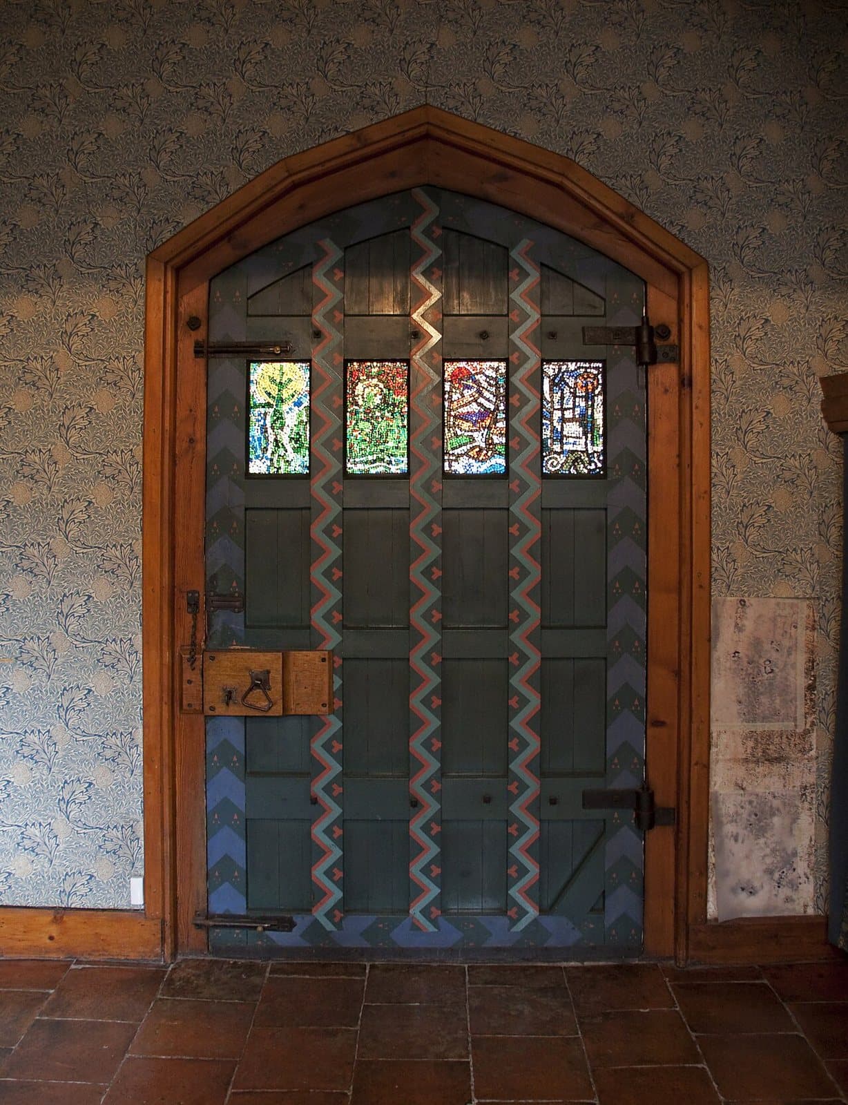 A dark green boarded door under a pointed wooden frame, painted with zigzag bands, with four small stained-glass panels and iron strap hinges