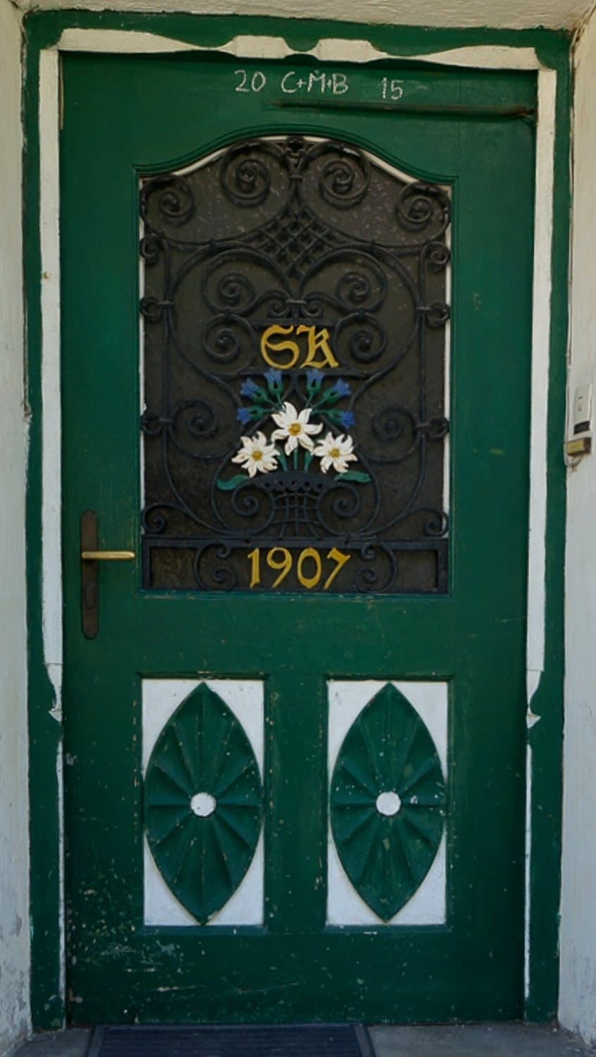 A green-painted wooden door with a scrolled iron grille over the glazing, painted edelweiss, the date 1907 and two pointed oval lower panels