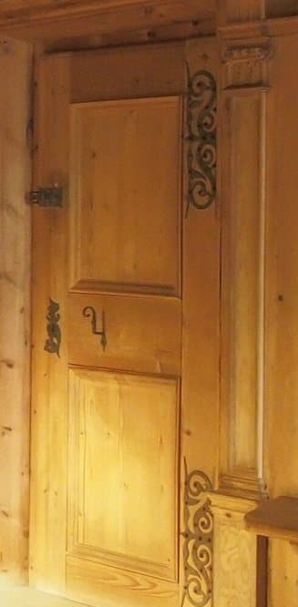 A pale two-panel wooden door in a paneled Tyrolean room, with scrolled black iron strap hinges and a carved pilaster beside it