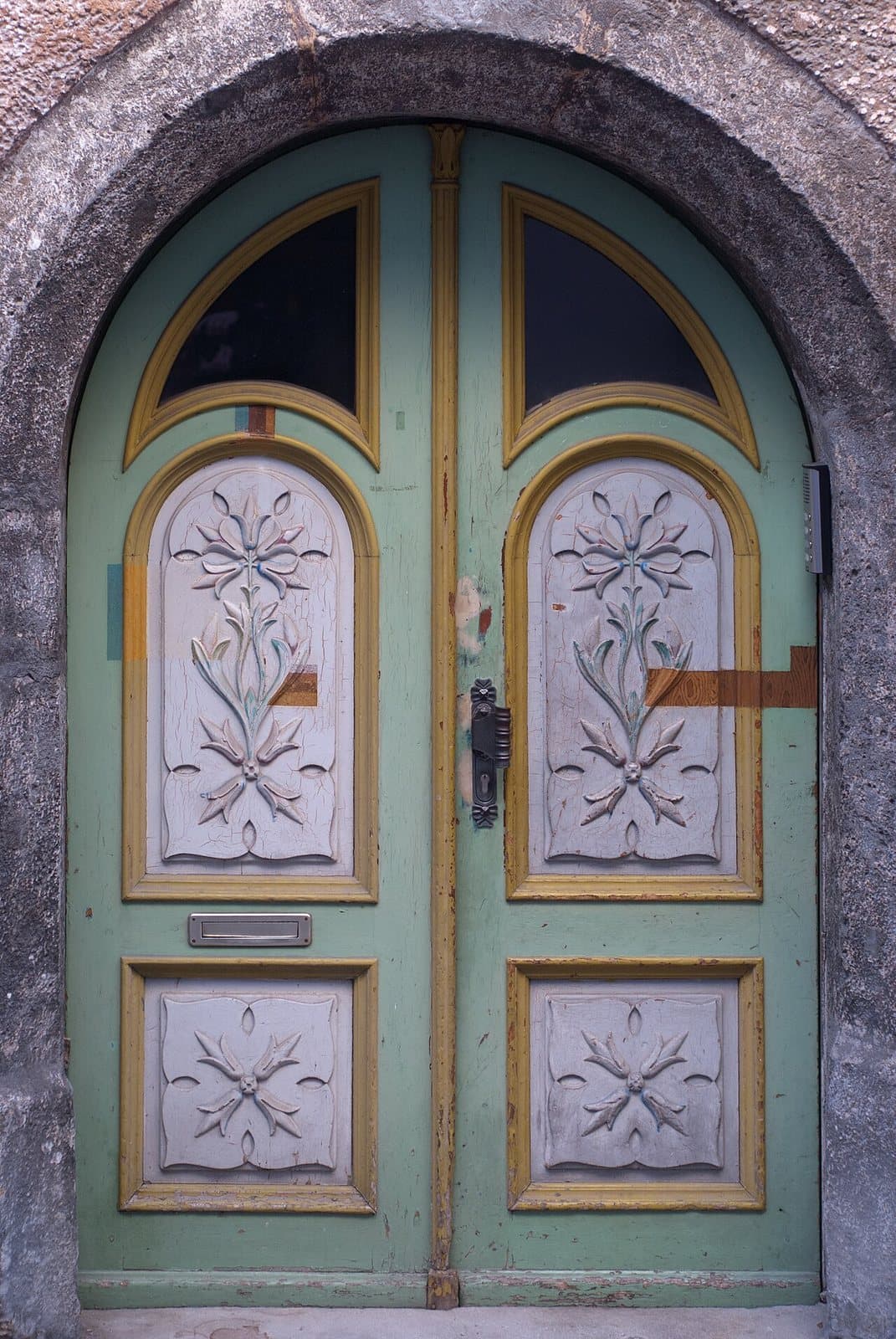 Arched double doors painted green and ochre, with glazed top lights and pale panels carved with stylized flowers, in a stone archway