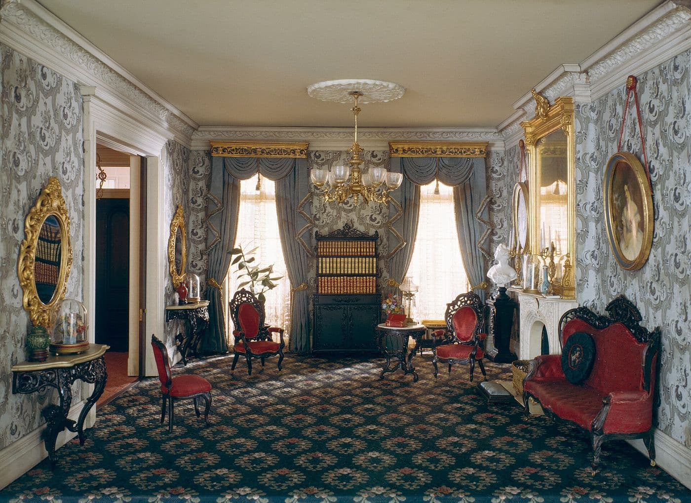 Miniature Victorian parlor with tall draped windows, a central built-in bookcase and red upholstered chairs
